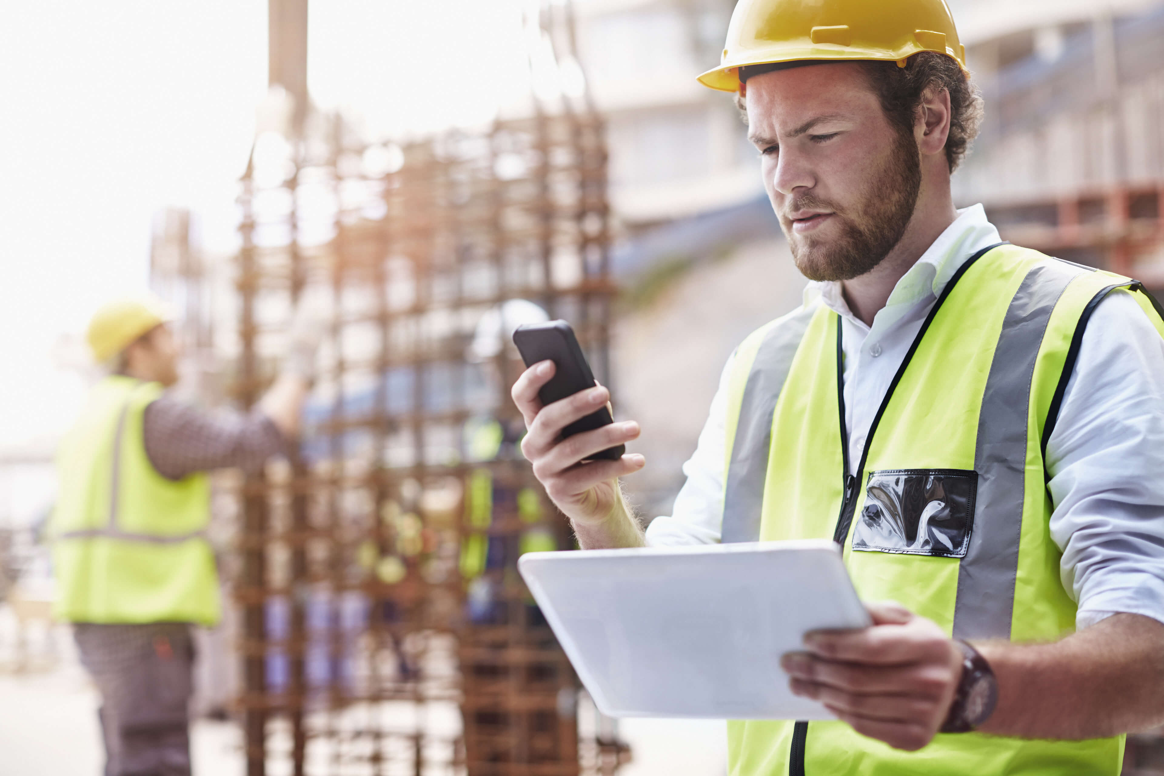 Bauarbeiter auf einer Baustelle der ein Tablet in der einen Hand hält und mit der anderen ein Smartphone bedient
